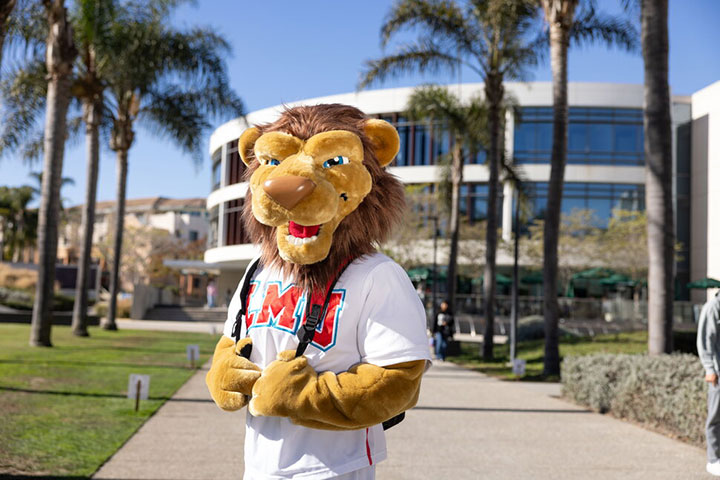 LMU mascot Iggy the Lion wearing a backpack with the William H Hannon Library in the background